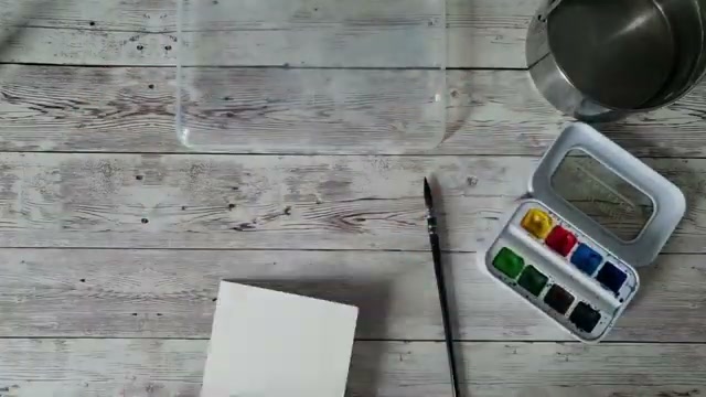 Présentation du couvercle de boîte de chocolat utilisé comme support, avec le matériel disposé sur la table : vaporisateur, pinceaux et godets d'aquarelle